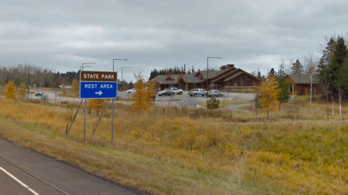 A sign for a state park rest area with a parking lot and rustic buildings in a grassy landscape under a cloudy sky.